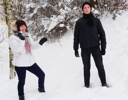 An elderly mother in a white jacket, in a scarf, holds a snowball in her hands near her son in black winter clothes against the backdrop of snow.