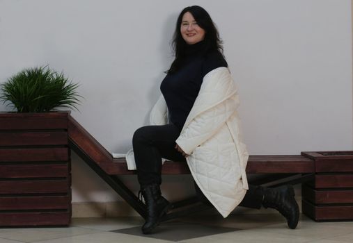 A pretty brunette plus-size girl in a black sweater and an unbuttoned white coat sits on a bench against a white wall.
