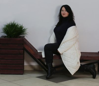 A pretty brunette plus-size girl in a black sweater and an unbuttoned white coat sits on a bench against a white wall.