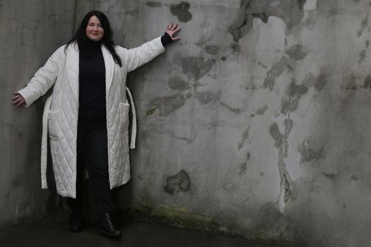 Portrait of a woman plus size model with dark hair in a white XXL winter coat and black trousers posing against wall.