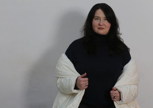 Large and gorgeous brunette woman. Pretty brunette woman in black sweater and white coat against a light wall looking at the camera.