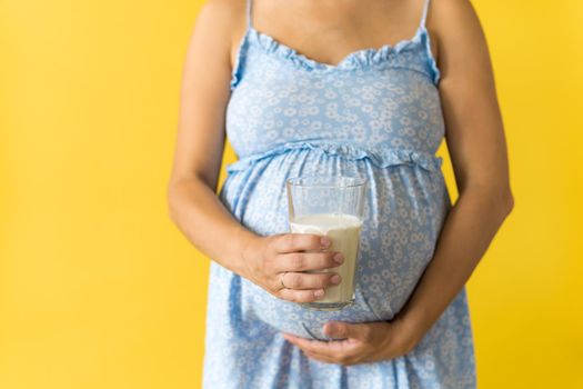 Motherhood, femininity, vitamins, calcium, healthy eating food, dairy, hot summer - croped portrait pregnant unrecognizable woman in floral blue dress holds drink glass of milk on yellow background.