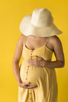 Motherhood, femininity, fashion,, hot summer - croped Close-up unrecognizable pregnant woman lady in beautiful vintage dress Provence straw hat with hands over tummy rubs tummy on yellow background.