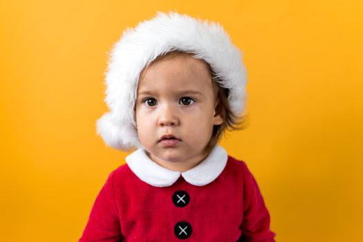 Serious Portraite Cute Happy Cheerful Chubby Baby Girl in Santa Hat Looking On Camera At Yellow Background. Child Play Christmas Scene Celebrating Birthday. Kid Have Fun Spend New Year Time Copy Space.