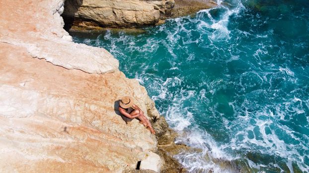 Beautiful girl sitting on a high rock and looking out to sea. top view. Girl in Hat on the edge of cliff. blue sea and high cliffs. Seascape. Girl at sunset. Sea tour. Blue sea, rocks