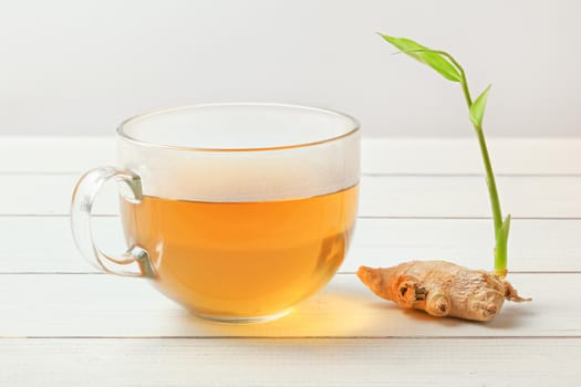 Transparent cup with hot tea, wet from steam on glass, dry ginger root with green sprout next to it.