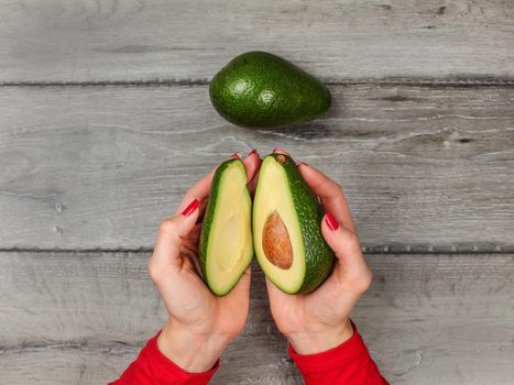 Tabletop view, young woman hands holding two halves of avocado cut in half, over gray wood desk, whole green pear next to it.