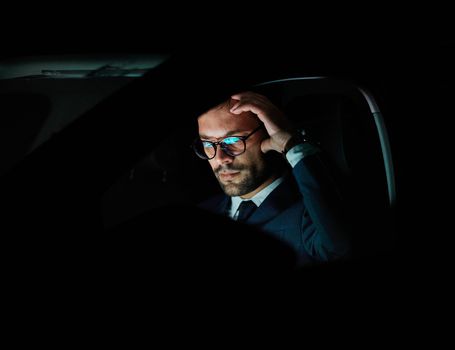 Portrait of a young businessman with tablet or laptop working late at night in office or looking at a device screen in car