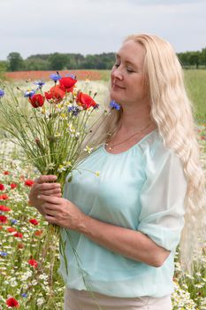 a beautiful middle-aged blonde woman stands among a flowering field of poppy, daisies, Cornflowers, and holds a bouquet of wild flowers and laughs. High quality photo