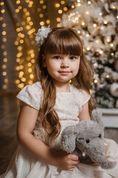 A girl in a festive light dress with a plush toy in her hands next to a Christmas tree, lights of garlands in the background. The concept of New Year holidays.