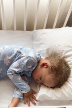 top view Cute little 2-3 years preschool baby boyl kid sleeping sweetly in white crib during lunch rest time in blue pajama with pillow at home. Childhood, leisure, comfort, medicine, health concept.