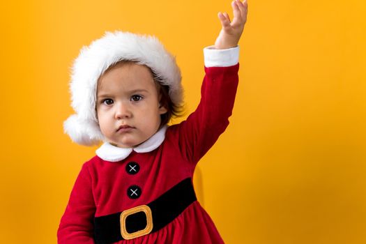 Serious Portraite Cute Happy Cheerful Chubby Baby Girl in Santa Hat Waving Hand Up At Yellow Background. Child Play Christmas Scene Celebrating Birthday. Kid Have Fun Spend New Year Time Copy Space.