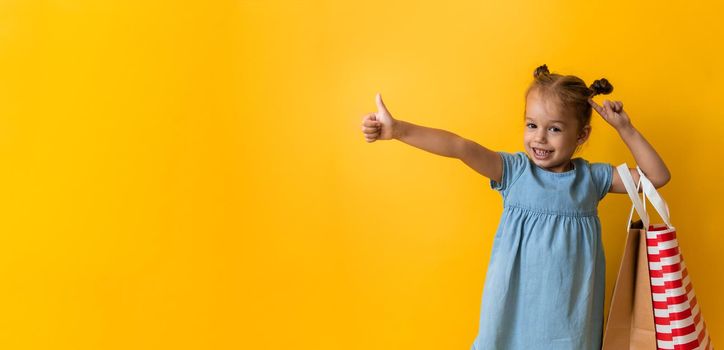 Banner Portrait Caucasian Beautiful Happy Little Preschool Girl Smiling Cheerful And Holding Cardboard Bags Thumb Up On Orange Yellow Background. Happiness, Consumerism, Sale People shopping Concept.