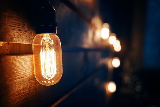 Antique incandescent Edison type bulb on wooden texture wall. Blurred warm garland light on background. Christmas night party mood. Holiday interior decorating. Copy space for congratulations.