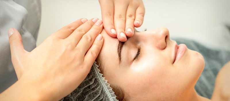 Young caucasian woman receiving facial massage by beautician's hands in spa medical salon