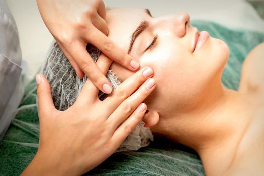 Young caucasian woman receiving facial massage by beautician's hands in spa medical salon