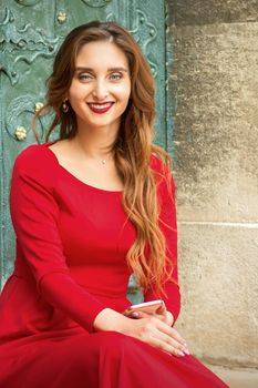 Portrait of a beautiful young caucasian traveling woman sitting at the door with smartphone smiling and looking at the camera outdoors