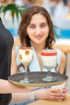 A young waitress carrying a tray with two different desserts in a two glass on the background of a young female client