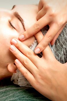 Young caucasian woman receiving facial massage by beautician's hands in spa medical salon