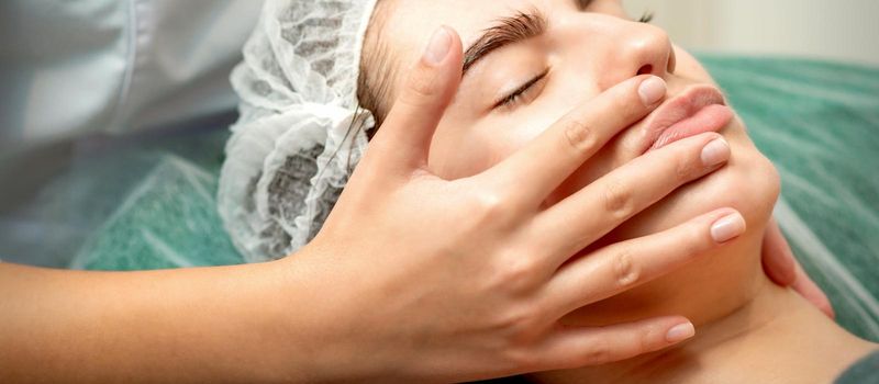 Young caucasian woman receiving facial massage by beautician's hands in spa medical salon