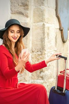 Beautiful young caucasian traveling woman in black hat looking on the smartphone smiling and sitting near the building on the street outdoors