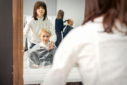 The female hairdresser puts on a transparent cellophane cape to the female client in a beauty hair salon