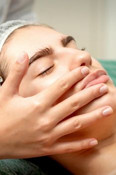 Young caucasian woman receiving facial massage by beautician's hands in spa medical salon