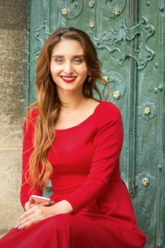 Portrait of a beautiful young caucasian traveling woman sitting at the door with smartphone smiling and looking at the camera outdoors