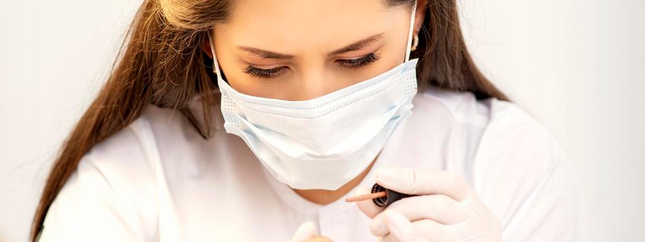 Manicure master in the white protective mask is painting nails with nail polish in a nail salon