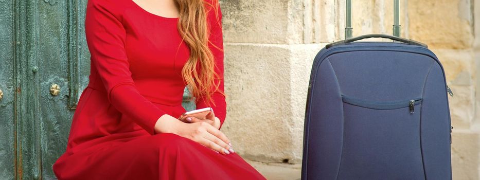Portrait of a beautiful young caucasian traveling woman sitting at the door with suitcase smiling and looking at the camera outdoors