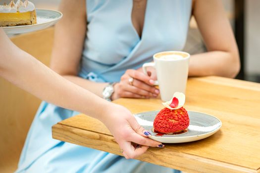 The waiter's hand puts on the table a plate with a red cupcake on the background of a female client in a cafe