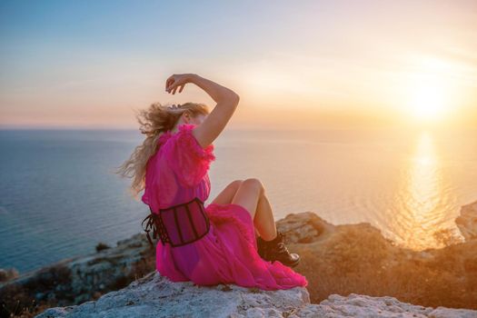 Side view a Young beautiful sensual woman in a red long dress posing on a volcanic rock high above the sea during sunset. Girl on the nature on blue sky background. Fashion photo