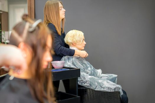The female hairdresser puts on a transparent cellophane cape to the female client in a beauty hair salon