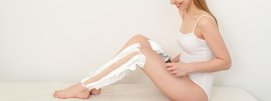 Beautiful young funny caucasian woman shaving legs with knife sitting on a white background