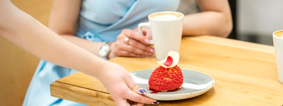 The waiter's hand puts on the table a plate with a red cupcake on the background of a female client in a cafe