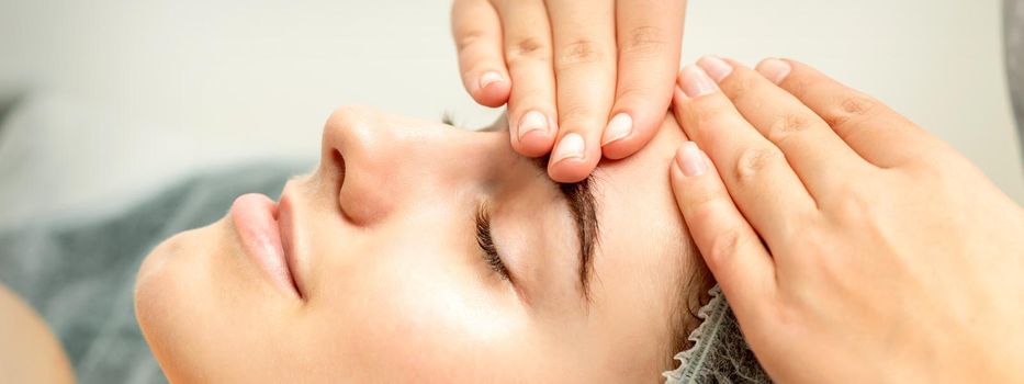 Young caucasian woman receiving facial massage by beautician's hands in spa medical salon