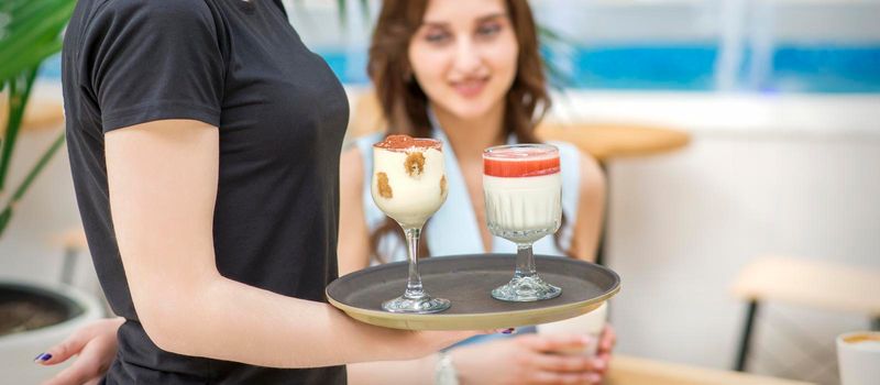 A young waitress carrying a tray with two different desserts in a two glass on the background of a young female client