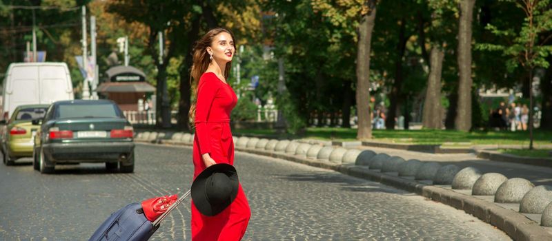 Beautiful young caucasian tourist woman with a suitcase in red long dress crosses the road through a crosswalk on the city street outdoors