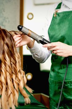 Back view of female hairdresser's hands curling women's hair with curling iron in a hair salon