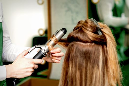 Back view of female hairdresser's hands curling women's hair with curling iron in a hair salon