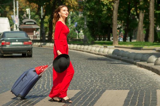 Beautiful young caucasian tourist woman with a suitcase in red long dress crosses the road through a crosswalk on the city street outdoors