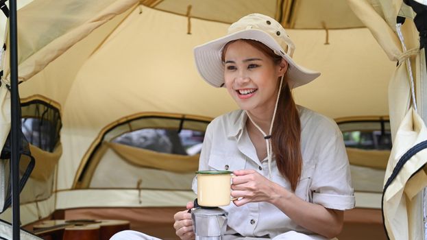 Pleasant asian woman sitting inside camp tent and drinking coffee. Travel, adventure and vacation concept.