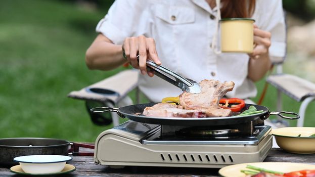 Young woman grilled bbq and preparing food for dinner during camping in nature park on beautiful day.