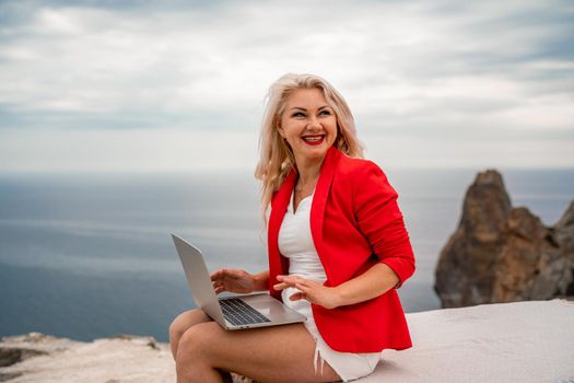 A woman is typing on a laptop keyboard on a terrace with a beautiful sea view. Freelancing, digital nomad, travel and vacation concept