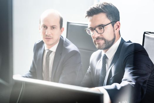 Image of two thoughtful businessmen looking at data on multiple computer screens, solving business issue at business meeting in modern corporate office. Business success concept.