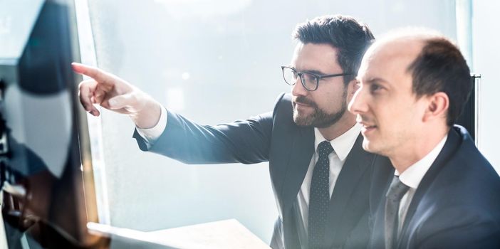 Image of two thoughtful businessmen looking at data on multiple computer screens, solving business issue at business meeting in modern corporate office. Business success concept.