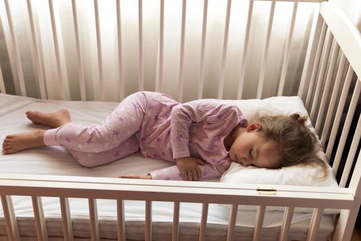 top view Cute little 3-4 years preschool baby girl kid sleeping sweetly in white crib during lunch rest time in pink pajama with pillow at home. Childhood, leisure, comfort, medicine, health concept.