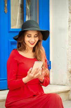 Beautiful young caucasian traveling woman in black hat looking on the smartphone smiling and sitting on stairs at the door outdoors