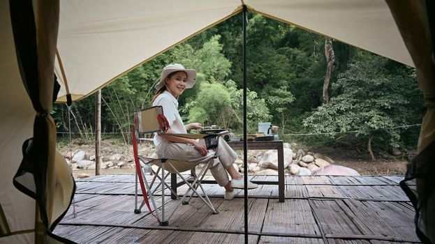 Peaceful asian woman sitting on folding chair near river bank during camping in nature park. Travel, adventure and vacation concept.