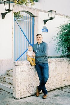 Dad hugs a baby sitting on a stone fence near the house. High quality photo